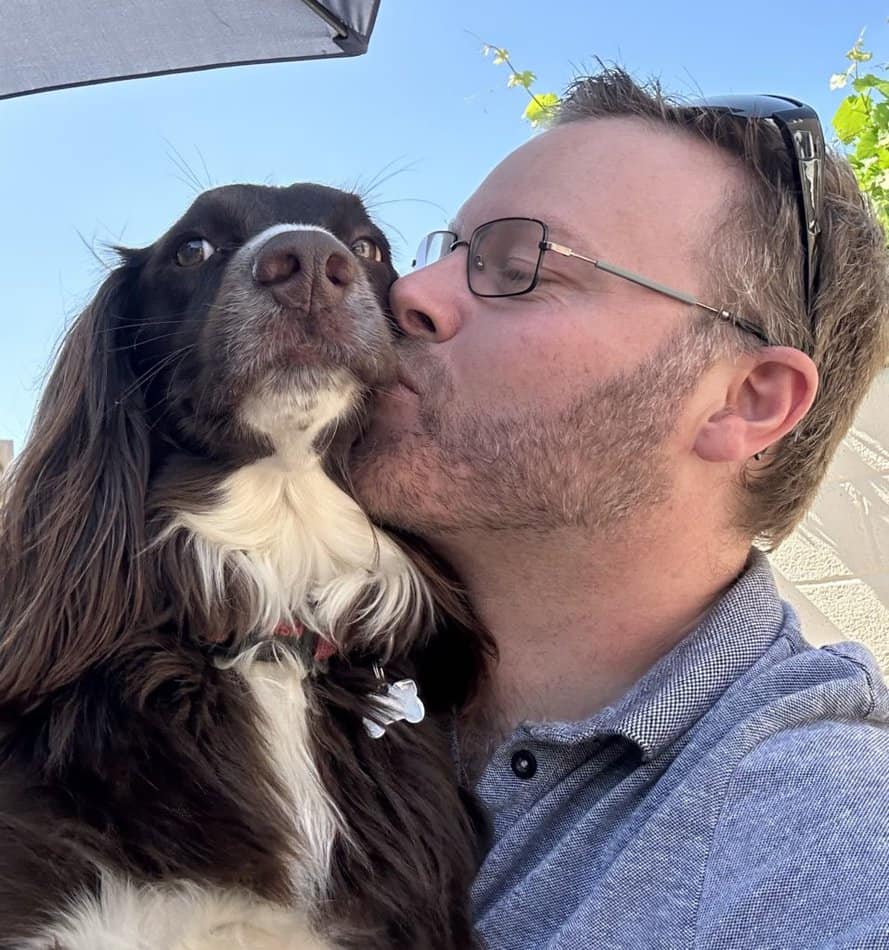 White male (Nick Sinclair) with blond hair and glasses, kissing brown sproker spaniel.  